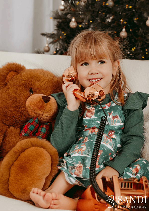 A young girl in an elegant Christmas setting, gently holding a festive gnome ornament during a photoshoot in The Evergreen Elegance set at Sandi Studio in Poundbury.
