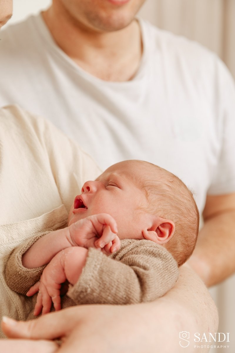 Profile view of a newborn baby resting on his mum's arm, wearing a brown knit cardigan.