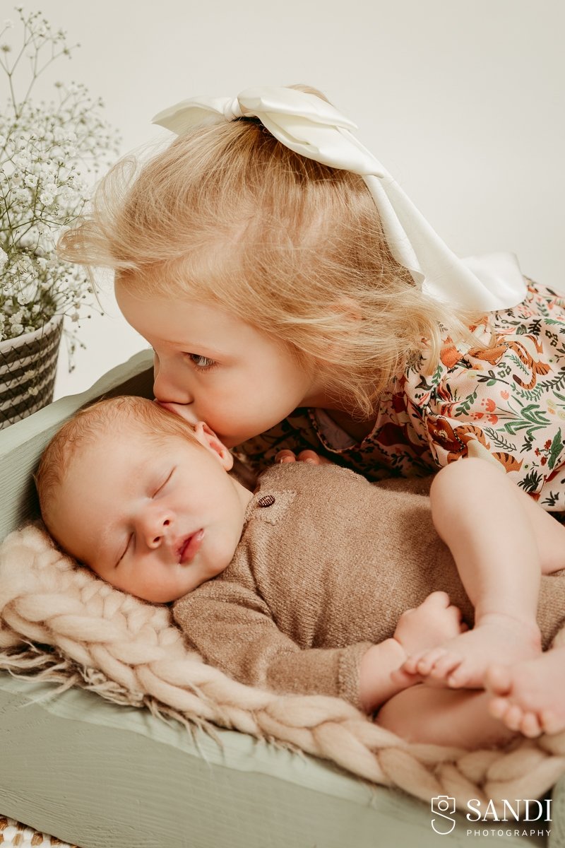 Close-up detail shot of a blonde toddler sister gently kissing her sleeping newborn brother on the forehead.
