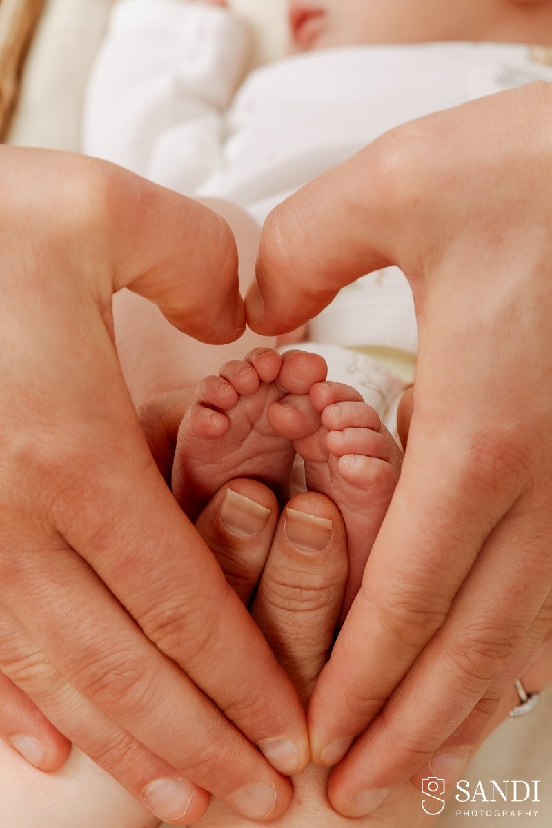 Close-up of parents' hands forming a heart shape around tiny newborn baby toes.