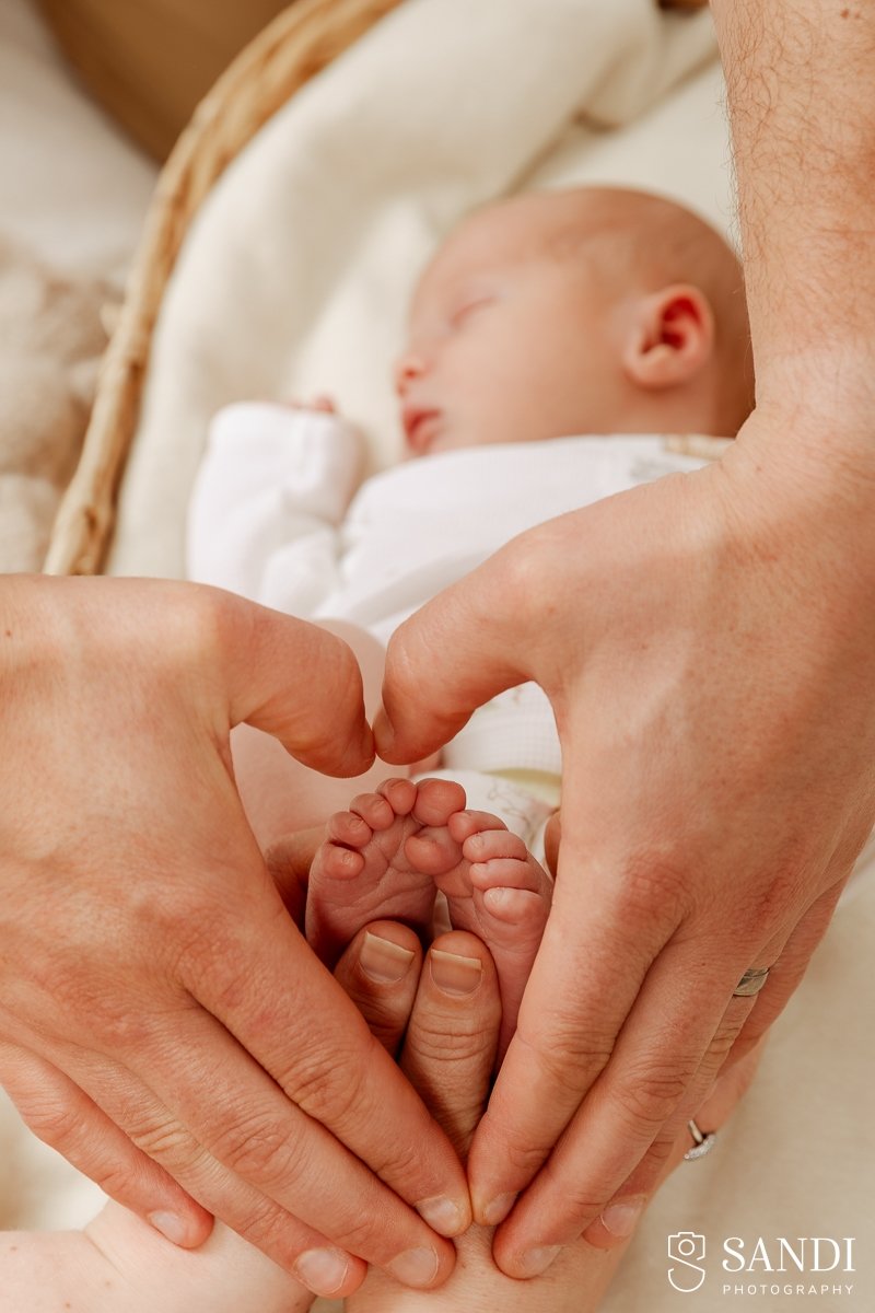 A view of a newborn baby lying on a white blanket with father's hands making a heart shape around the baby's feet.