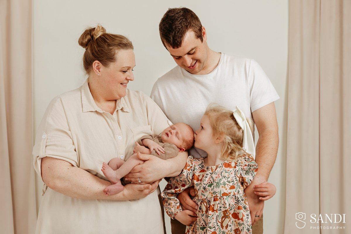Smiling young girl in a white lace dress sitting on a bed holding her newborn brother, with boho dried flowers in the background.