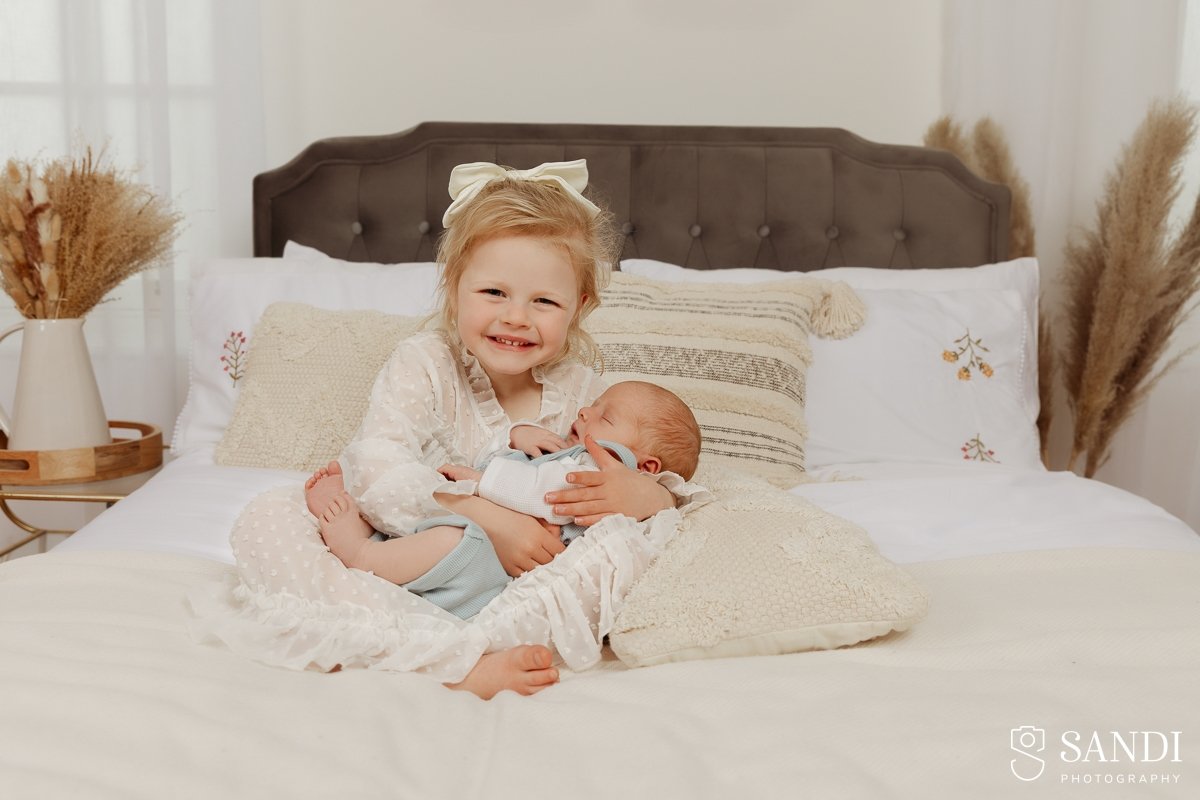 Smiling young girl in a white lace dress sitting on a bed holding her newborn brother, with boho dried flowers in the background.