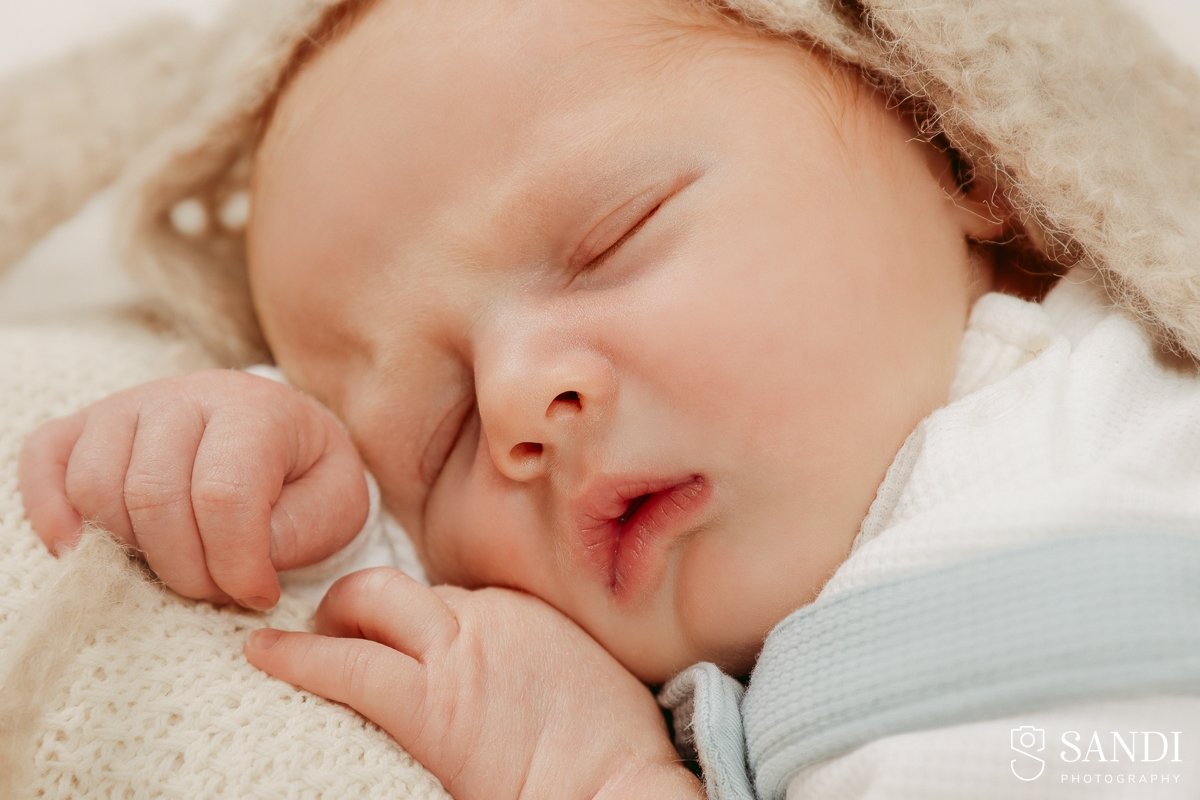 Extreme close-up of a sleeping newborn baby's face, showing details of eyelashes and lips, with a beige knit bonnet framing the face.