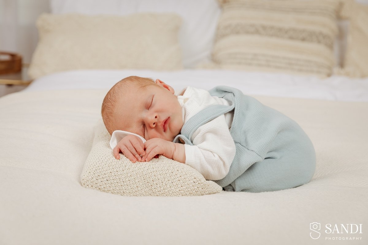 Newborn baby boy sleeping comfortably on his tummy on a cream blanket, wearing a light blue textured romper suit