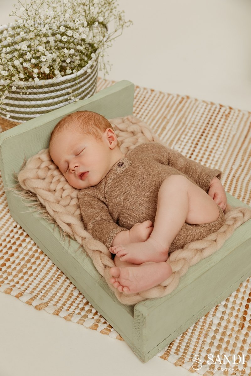 Newborn baby boy sleeping peacefully in a brown knit outfit on a sage green wooden bed with a chunky knit blanket.
