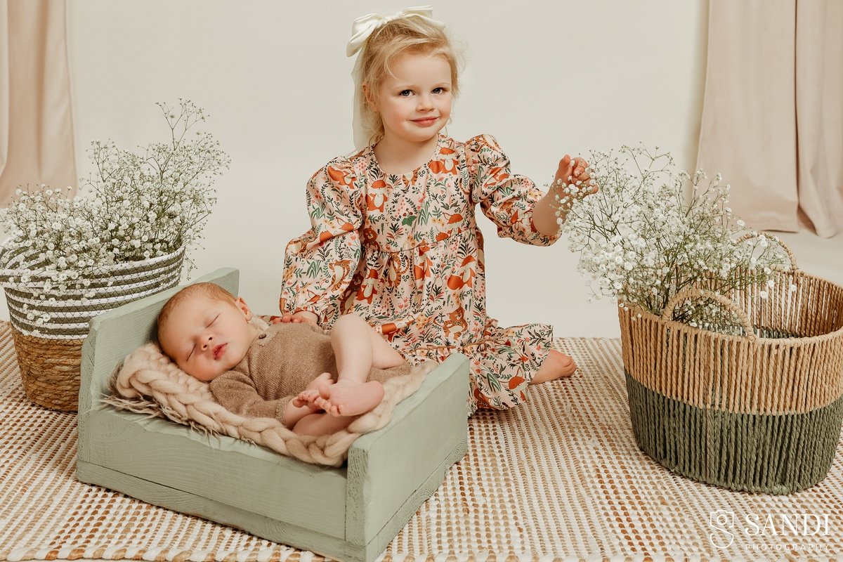 A young girl in a floral dress holding baby's breath flowers next to her newborn brother sleeping in a sage green wooden bed during a studio session.