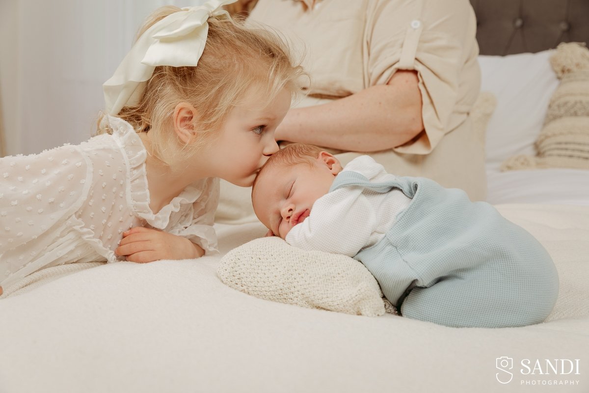 Blonde toddler sister gently kissing her sleeping baby brother on the forehead as he lies on a white textured pillow.